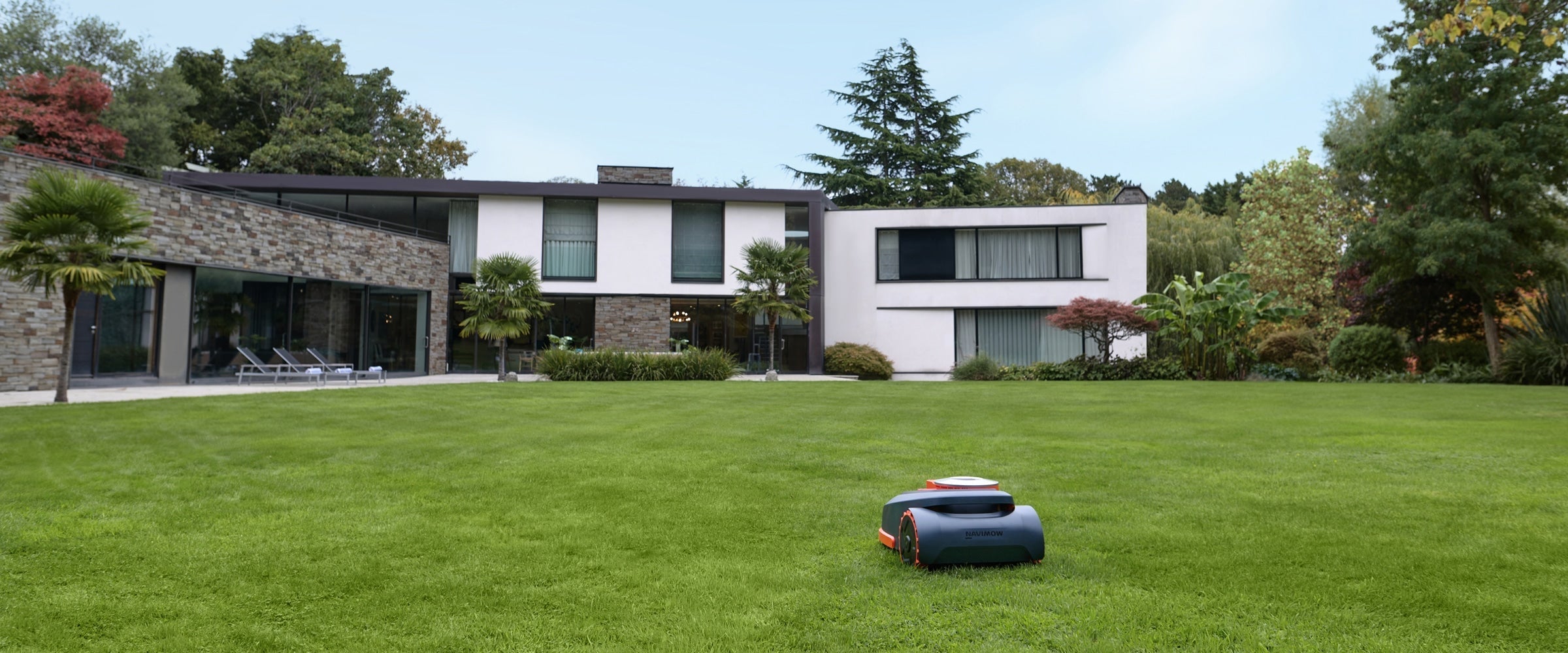 Segway Navimow H2 robot lawn mower crosses manicured turf near lounge chairs and a glass-walled house