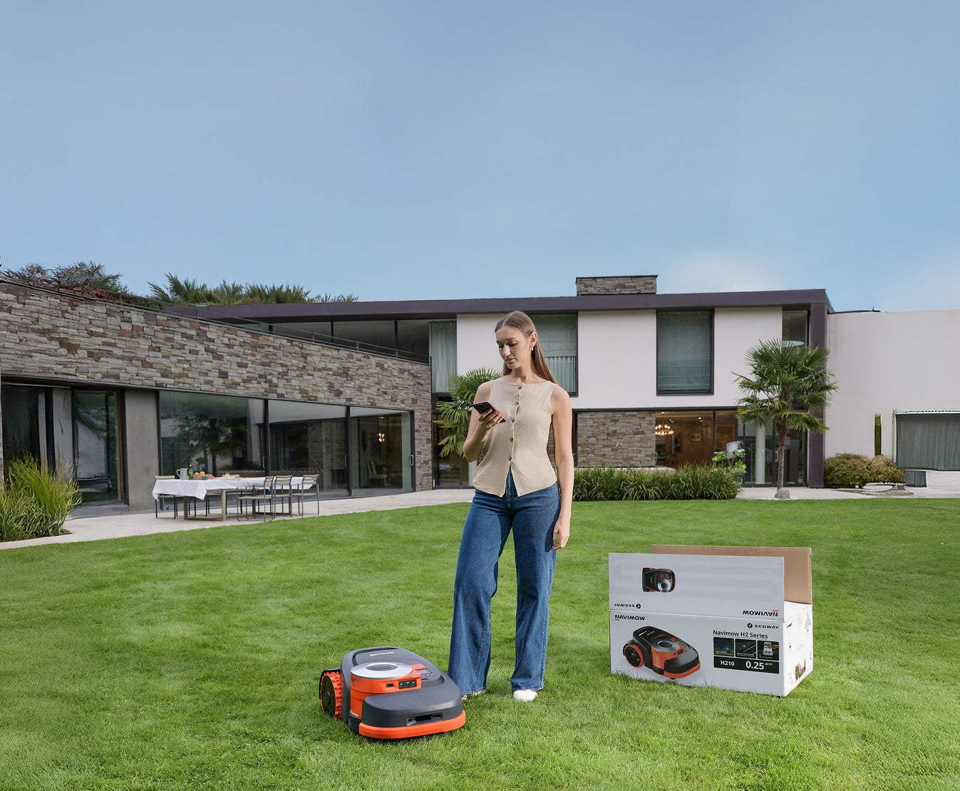 Segway Navimow H2 robot lawn mower and retail carton with woman on phone in a modern backyard